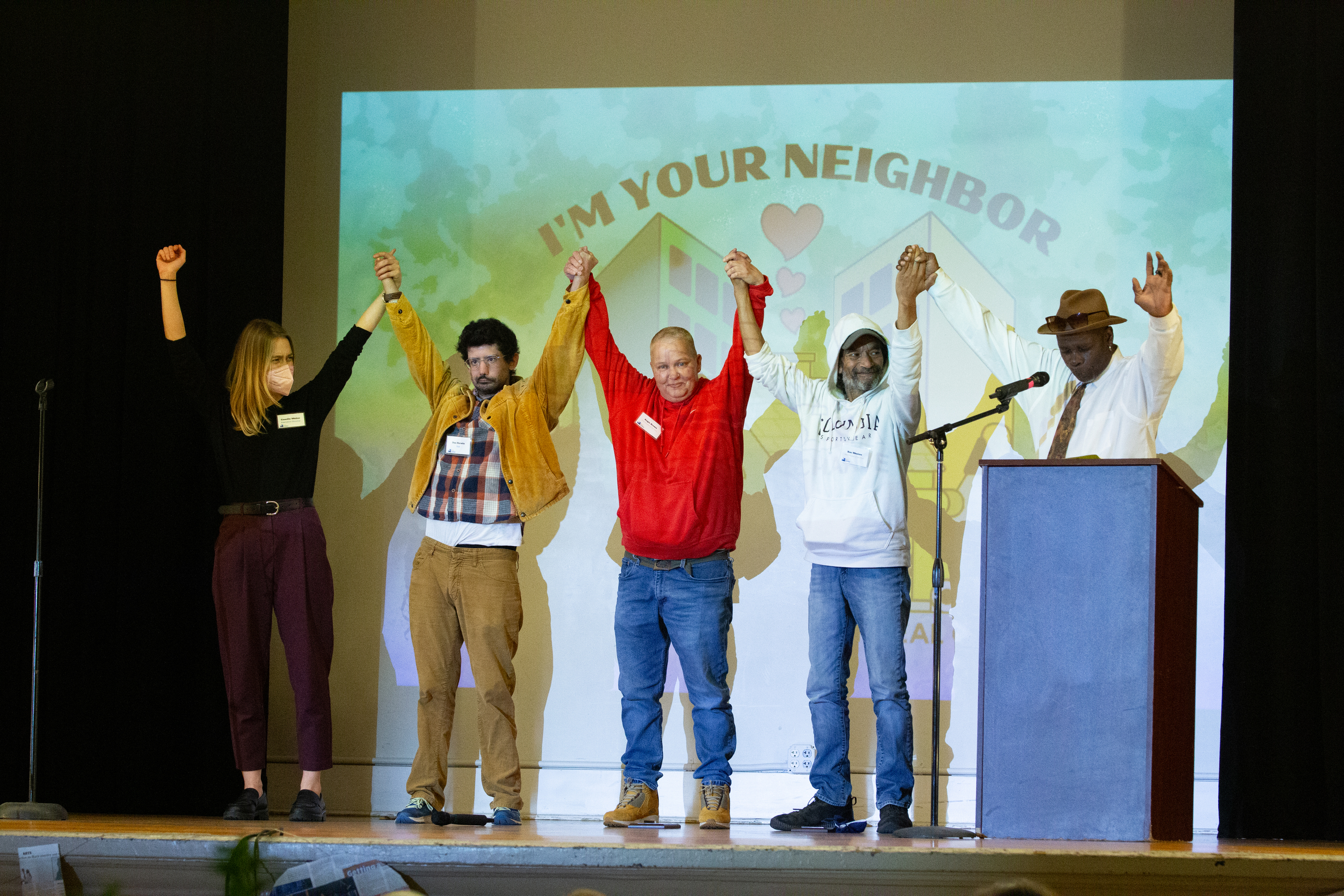 Real Change staff, board, event host, and award winners hold raised hands on stage at the 29th anniversary party.